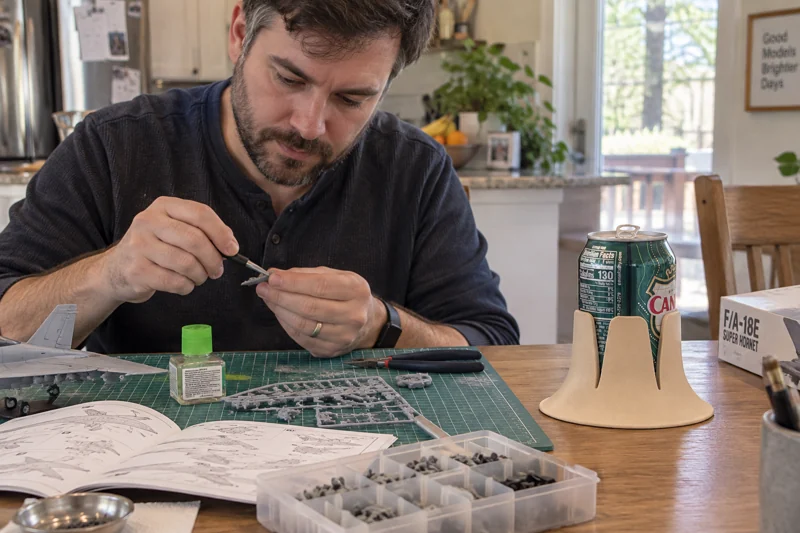 Adult working on hobby model kit at kitchen table with opened standard 12-ounce soda can in Steadi on clear tabletop spot outside hobby work mat. AI illustration.